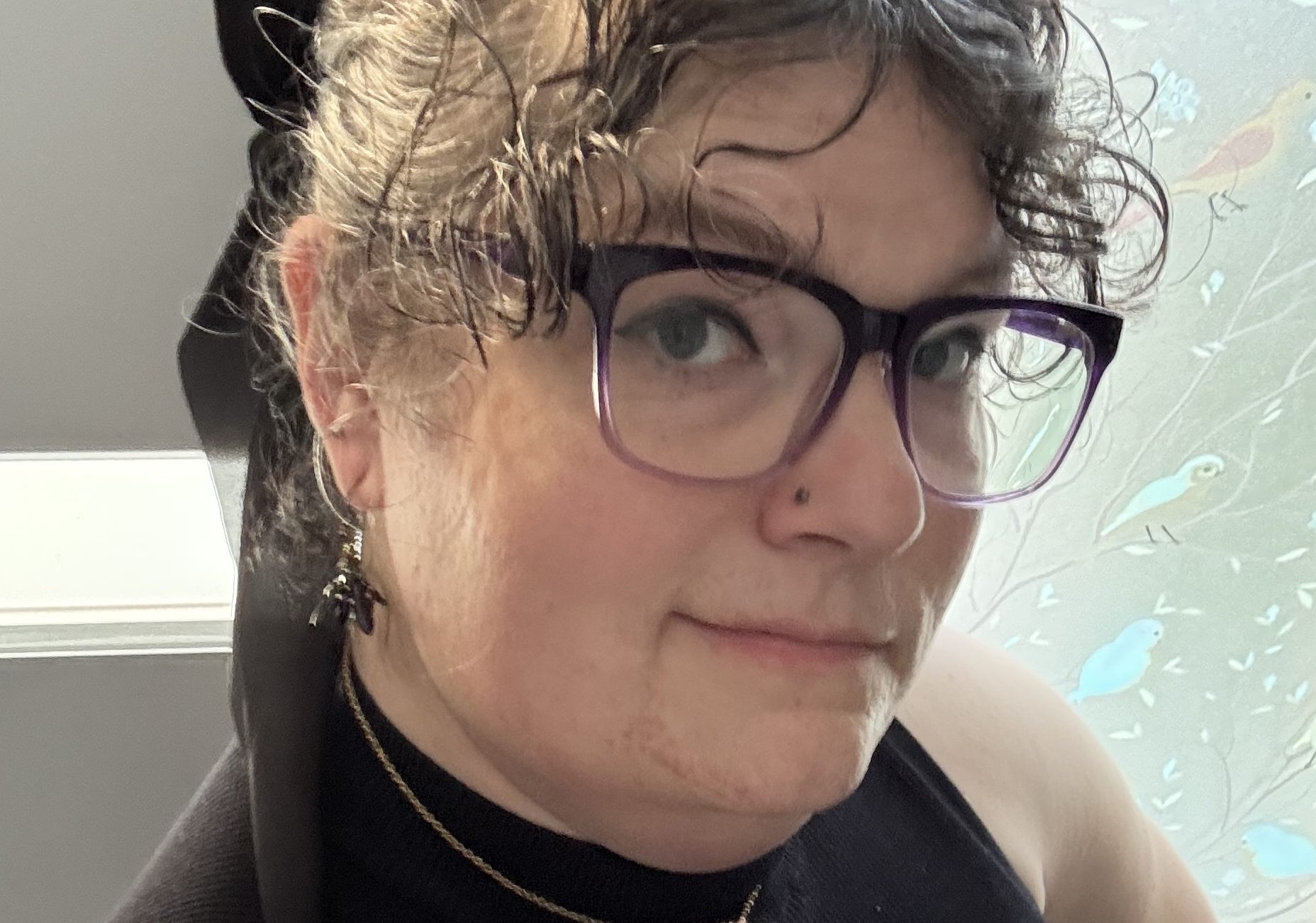 Selfie of a woman with curly dark hair, glasses, and a nose piercing. She has a bow in her hair and a sleeveless top.