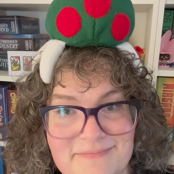 Photograph of Phoebe – a white woman with curly hair and glasses – smiling at the camera with a metroid plushie on her head.